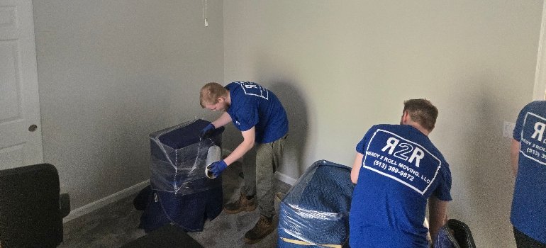 Packing Service 3 Two movers in blue RZR uniforms carefully wrap furniture in protective covers inside a sparsely furnished room.