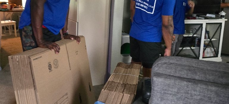 Moving Boxes 4 Three people wearing blue shirts stand indoors, holding large flat cardboard boxes and bundles of flattened moving boxes near a gray couch.
