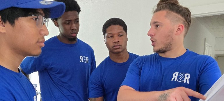 Residential Movers 2 A group of four individuals in matching blue t-shirts, engaged in conversation and collaboration in a bright, minimalistic space.