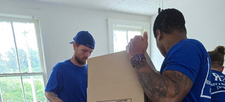 Residential Movers 3 Two movers in blue shirts are working together to lift a cardboard box inside a bright room with large windows.