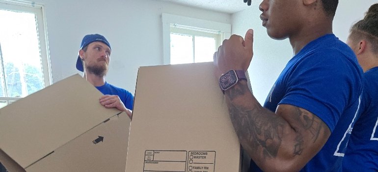 Moving Boxes 2 Two men in blue shirts carry moving boxes in a well-lit room with white walls and a window. The mood appears focused and collaborative.