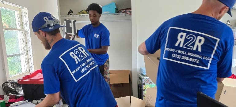 Local Movers 2 Three local movers in blue t-shirts with the "Ready 2 Roll Moving" logo, packing items into boxes in a cluttered room.
