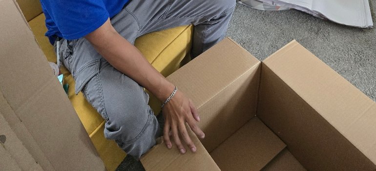Moving Boxes 3 A person in a blue shirt and gray pants sits on a yellow sofa, assembling a cardboard box on a gray carpet. The scene conveys focus and productivity.