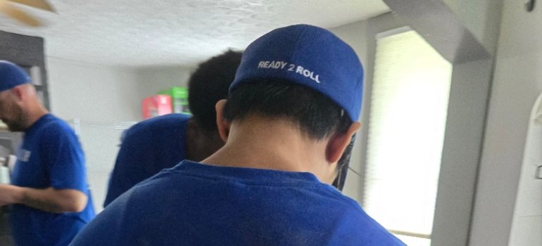 Piano Movers 2 Person wearing a blue cap with "READY 2 ROLL" text on the back, standing indoors with two others, all dressed in blue shirts.
