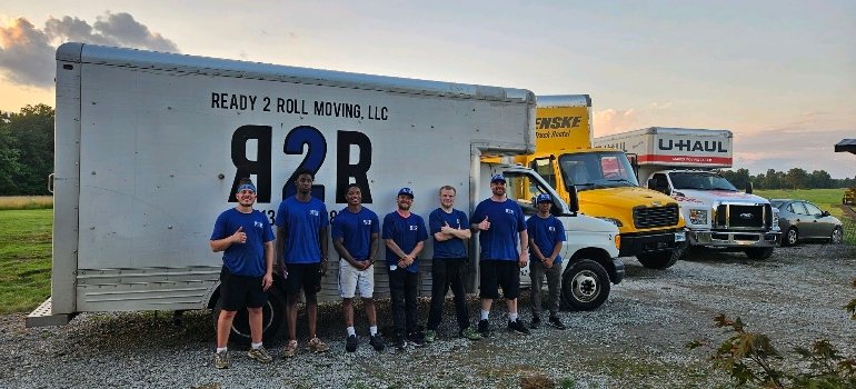 Residential Movers 1 A group of seven residential movers in blue shirts stands smiling in front of a "Ready 2 Roll Moving" truck, with rental trucks nearby.