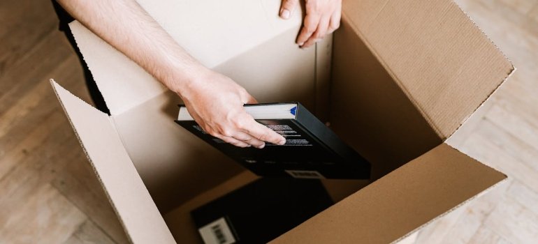 The Best Way to Pack Books for Moving 1 A person places a book into an open cardboard box, following tips for how to pack books for moving