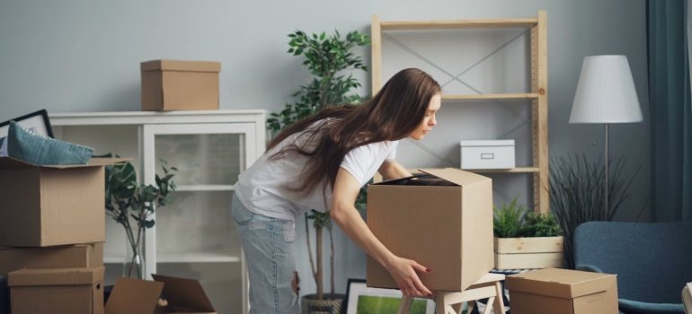How To Clean An Apartment Before Moving In 3 A person is unpacking boxes in a cozy, plant-filled living room, surrounded by cardboard boxes and minimalistic furniture.