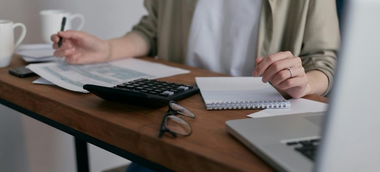 Moving Estimate vs Final Invoice: What’s the Difference 2 Person sitting at a desk with a calculator, notebook, and laptop while reviewing papers, illustrating how customers compare a moving estimate vs final invoice before planning their relocation costs.