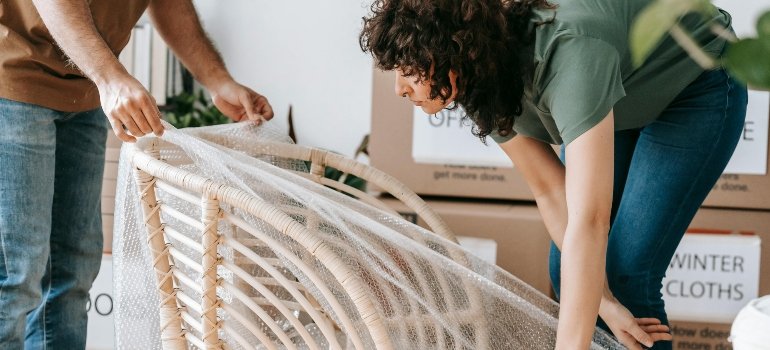 Preparing for movers: What to do the week before your move 2 Two people carefully wrapping a rattan chair in bubble wrap while organizing boxes labeled for winter clothes.