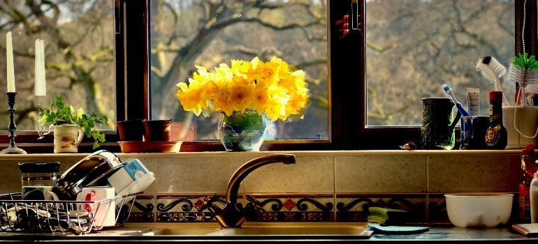 Practical Checklist of Things You Need When Moving Out 1 Sunlit kitchen window with a vase of yellow flowers and dishes on the counter.